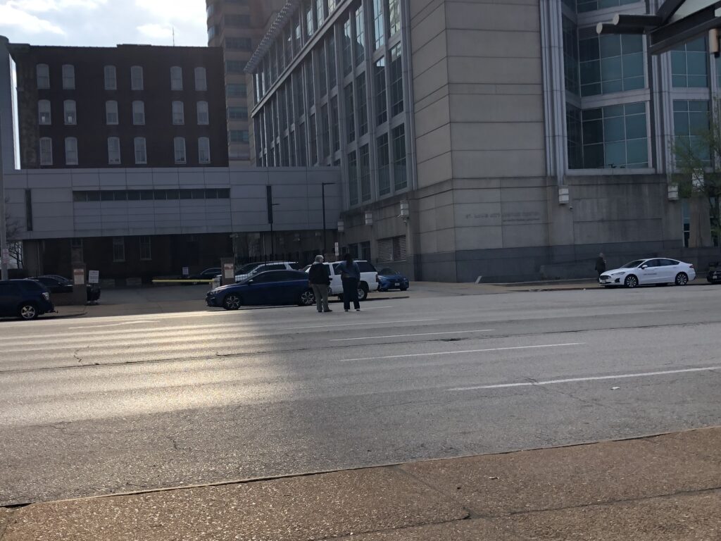 Pedestrians pause in the center of a wide street without a crosswalk, waiting for their chance to cross