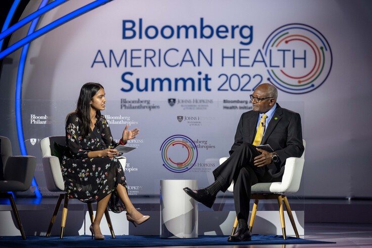 A woman on left speaking with a man, both seated, in front of a sign that reads
