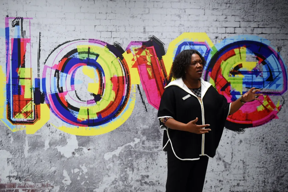 A woman with arms extended, speaking to an audience, standing in front of colorful street art that reads