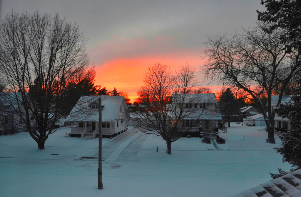 The sunsets behind two houses in a neighborhood covered in snow.