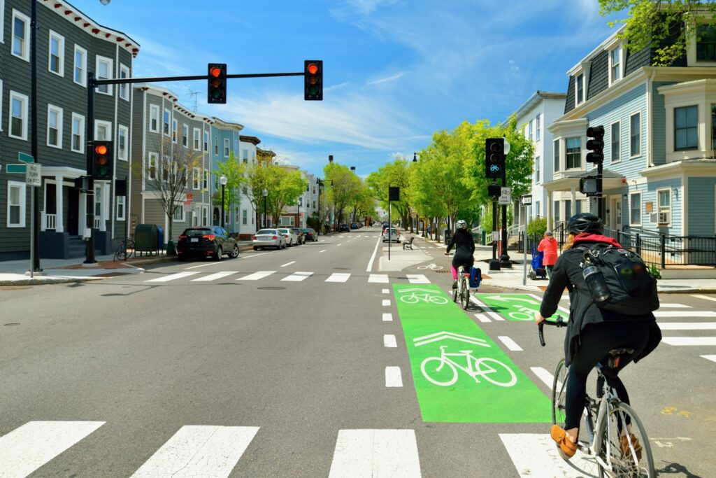 Two bikers cross a street while their bike lane signal is green. They are riding down a suburban street where houses line the streets on both sides, and the only cars visible are parked ones.
