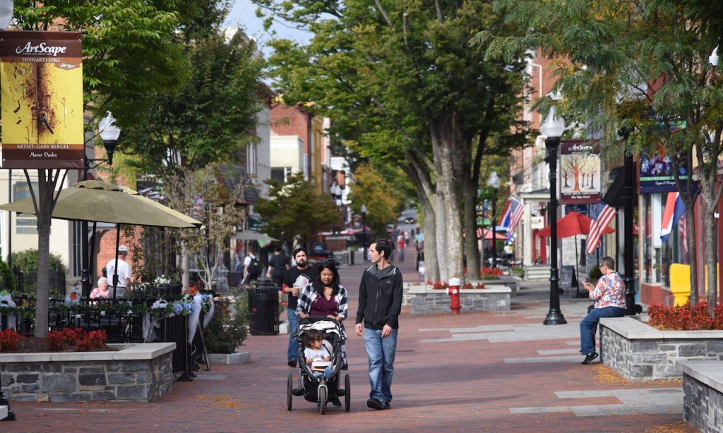 a young family with others pictured around the frame are walking through a downtown street with small shops and trees in planters