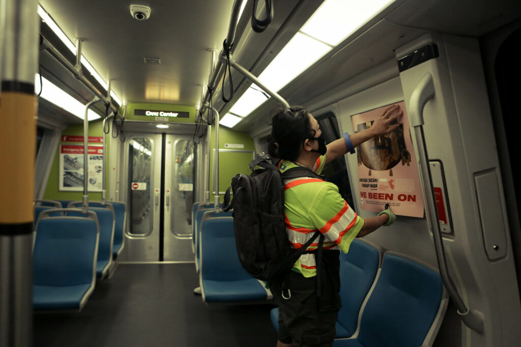 Image of person installing a pink poster featuring an image of a wooden cultural mask onto the side of a train car.