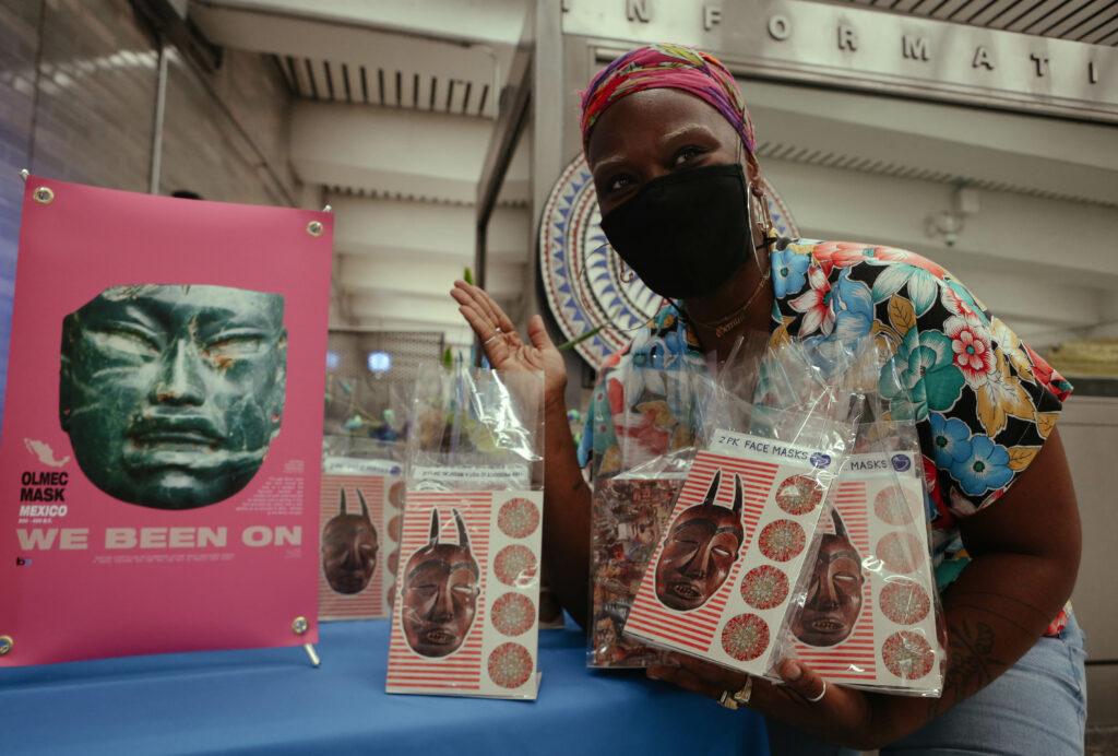 Image of person crouching next to a pink poster that reads “We been on” and features a slate colored cultural mask. Person holds in their hands a number of clear giveaway bags containing art postcards.