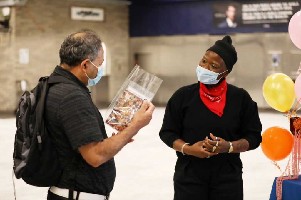 Image of two people wearing masks talking inside a transit station. Person on the left holds in their hand a clear plastic bag with a pink postcard inside.