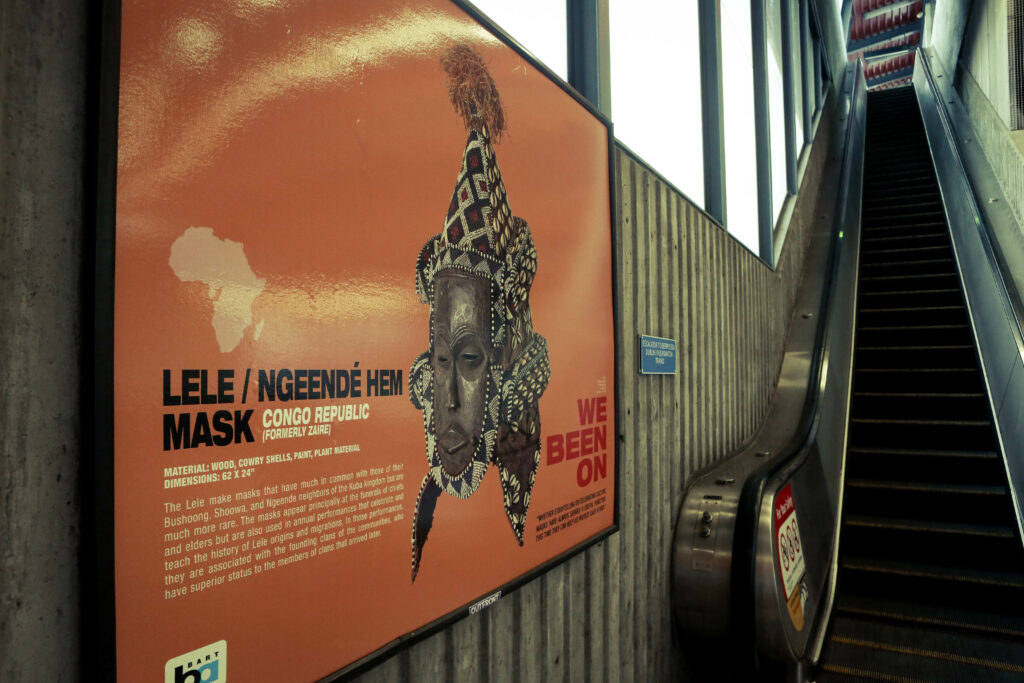 Image of a orange poster featuring a wooden mask from Congo Republic in front of an escalator in a transit station.