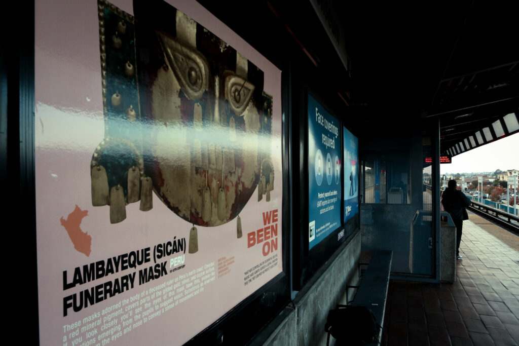 Image of a transit station waiting area. On the left is a light pink sign featuring a gold mask from Peru.