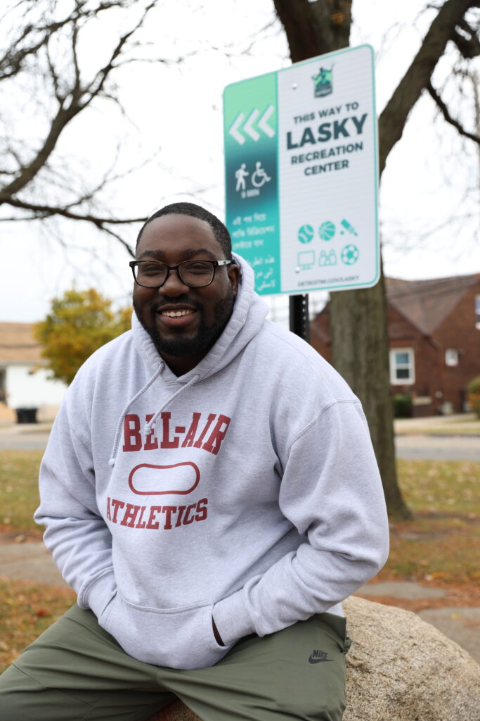 Person sits on a boulder in front of a teal and white wayfinding sign featuring the Detroit logo that directs people to nearby Lasky Recreation Center.