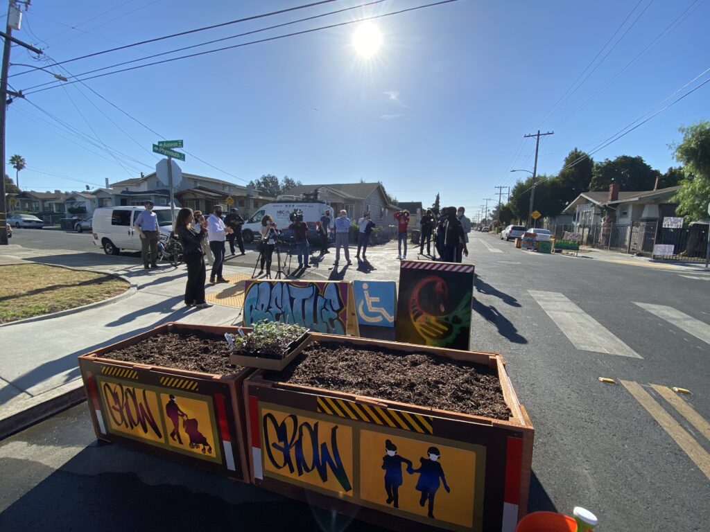 Image of planter barricades that are bright yellow and feature the words “GROW” and stenciled images of small children wearing masks and holding hands. In the background of the photo, people listen to a speaker.