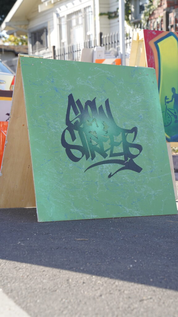 Image of green wooden sign with the words “Slow Streets.”