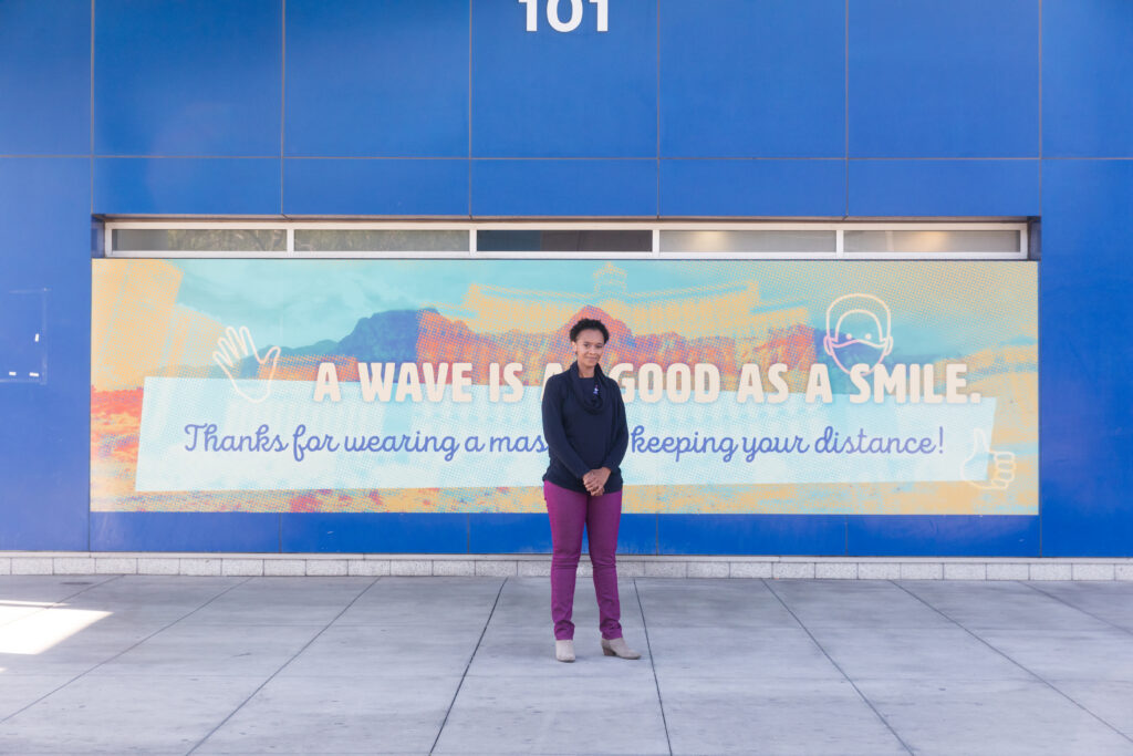 Image of a person standing in front of a sign on an exterior wall of Bonneville Transit Center which reads "A wave is as good as a smile."