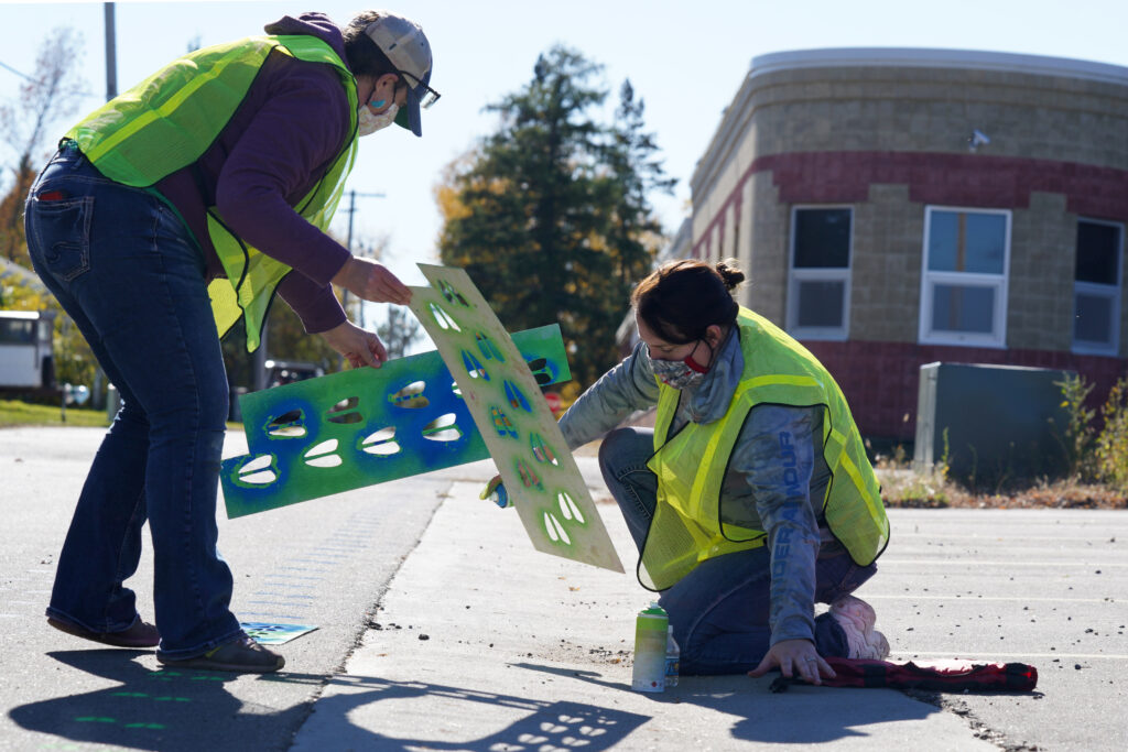 People use stencils to spray paint green animal footprints on a street.