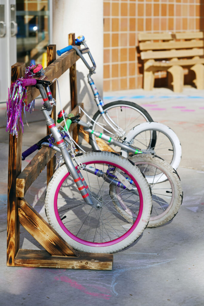 Image of pink, green, and white play sculptures made up of bicycle parts leaning against a fence.