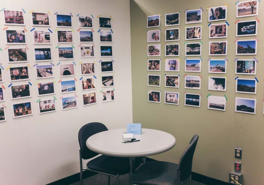 Image of the corner of a room with numerous photos taped to the walls. In the foreground is a table with two chairs.