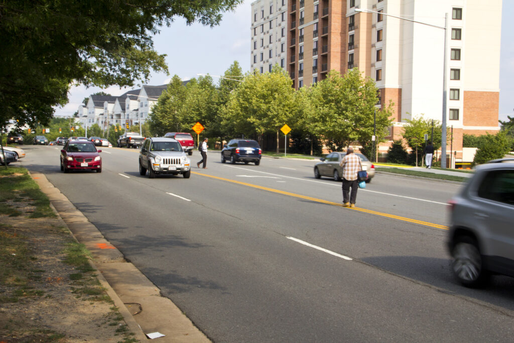 image of people crossing a five-lane street in the DC metro area where there are no sidewalks and crosswalks far apart near a metro station