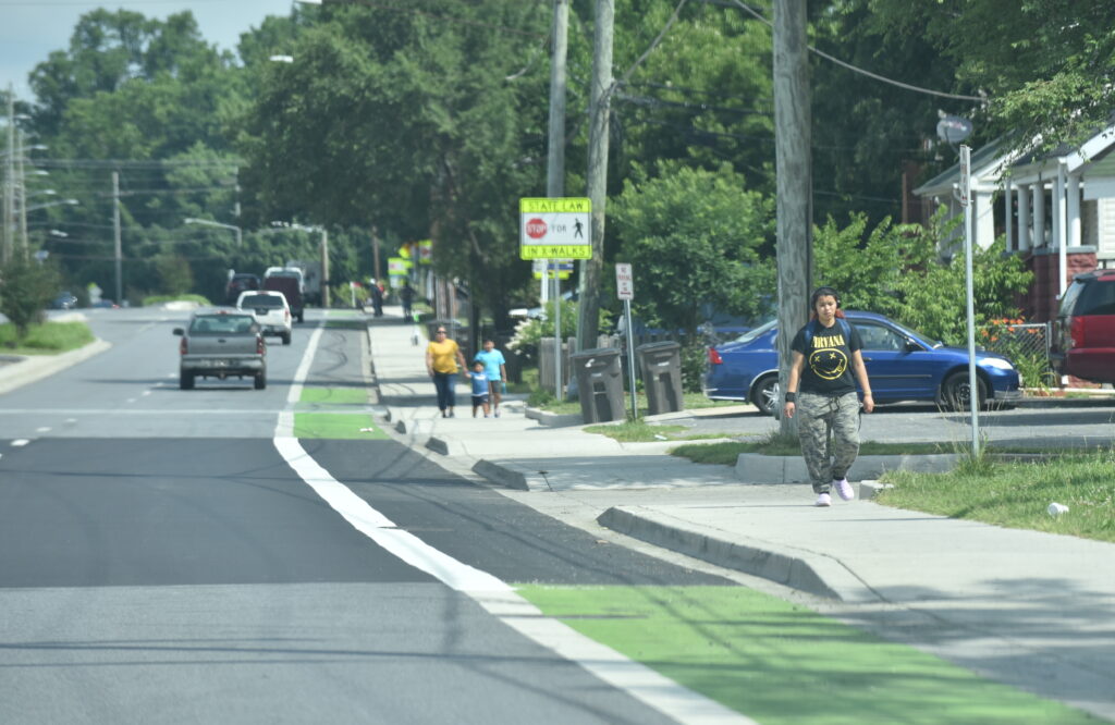 A photo of a street with two lanes headed away and a bike lane next to high speed traffic, where the sidewalk has many curb cuts and people are walking toward the camera