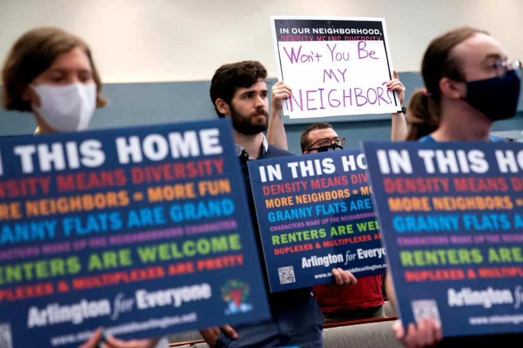 Three pro-housing advocates holding colorful signs at an Arlington Board meeting.