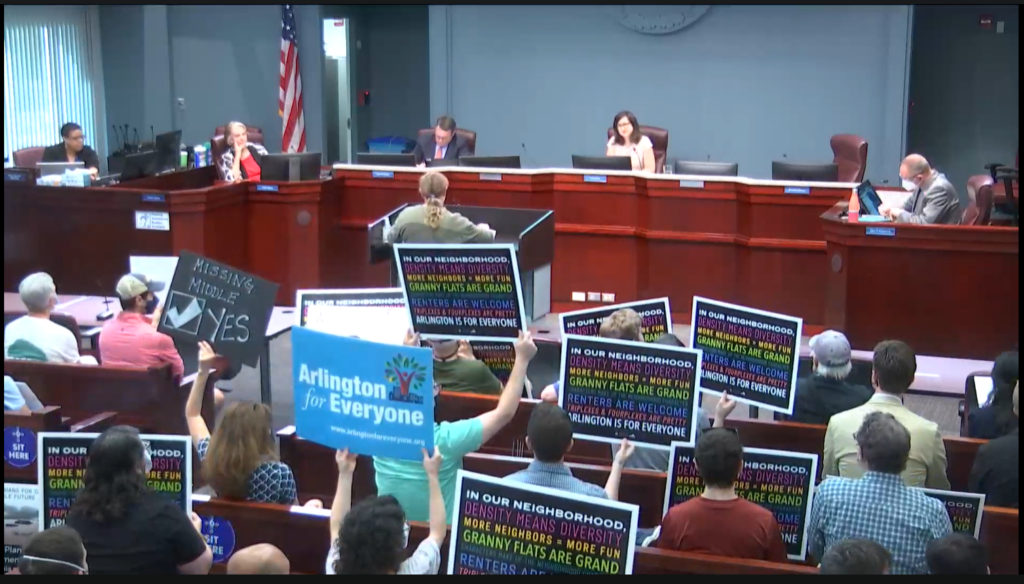 Pro-housing advocates with colorful signs attend an Arlington County Board Meeting, in a blue-painted room. In the center of the room is the board, sitting at a large wooden desk.