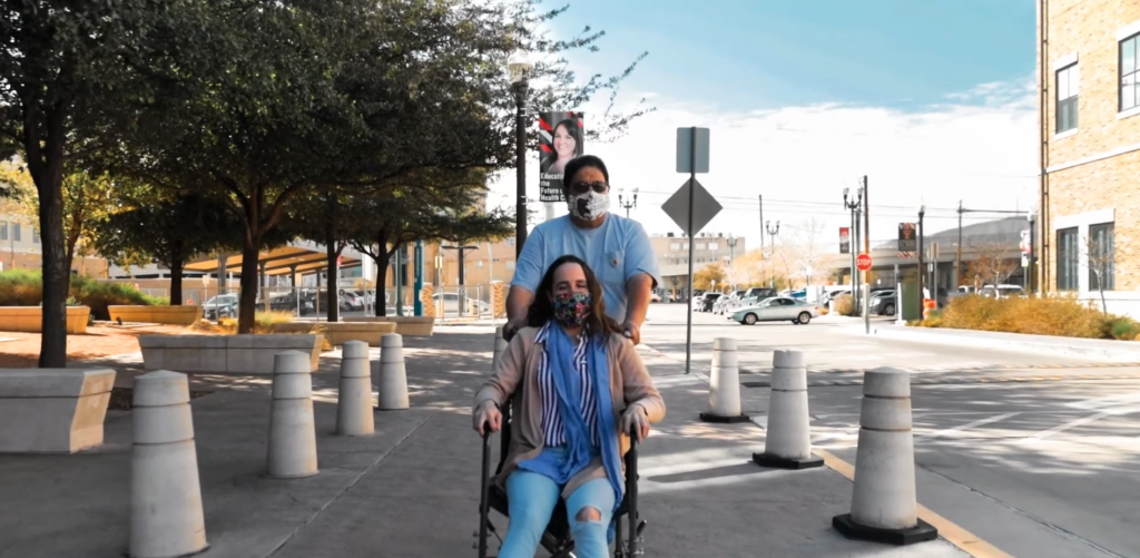 A man pushes a woman in a wheelchair inside a sidewalk protected by bollards