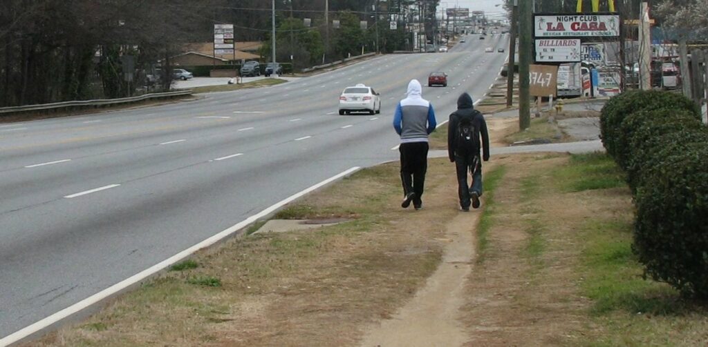 two people walk in a desire path tread into grass adjacent to a 7 lane arterial road in Atlanta