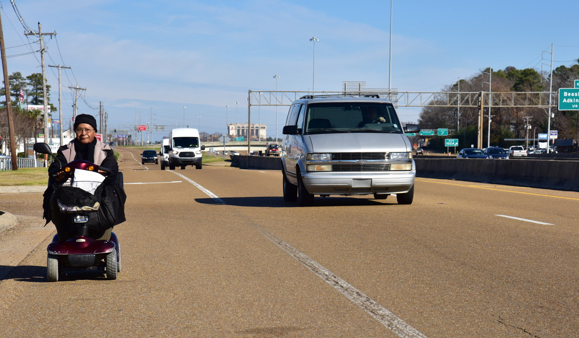 a woman in a wheelchair travels toward the camera in a shoulder of a highway with cars to the right of her.