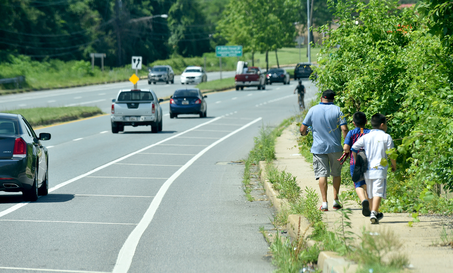 a family of three walks on an overgrown sidewalk next to a busy highway