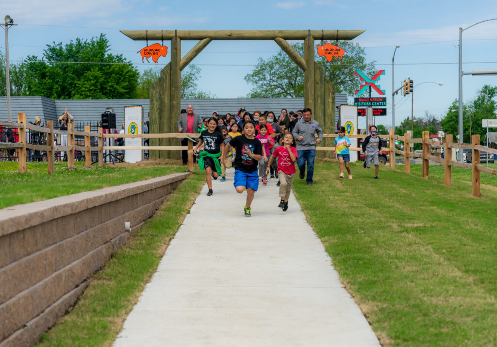 A large group of kids run toward the camera through a gateway entrance along a path with grass on both sides of the WahZhaZhe Heritage Park Trail