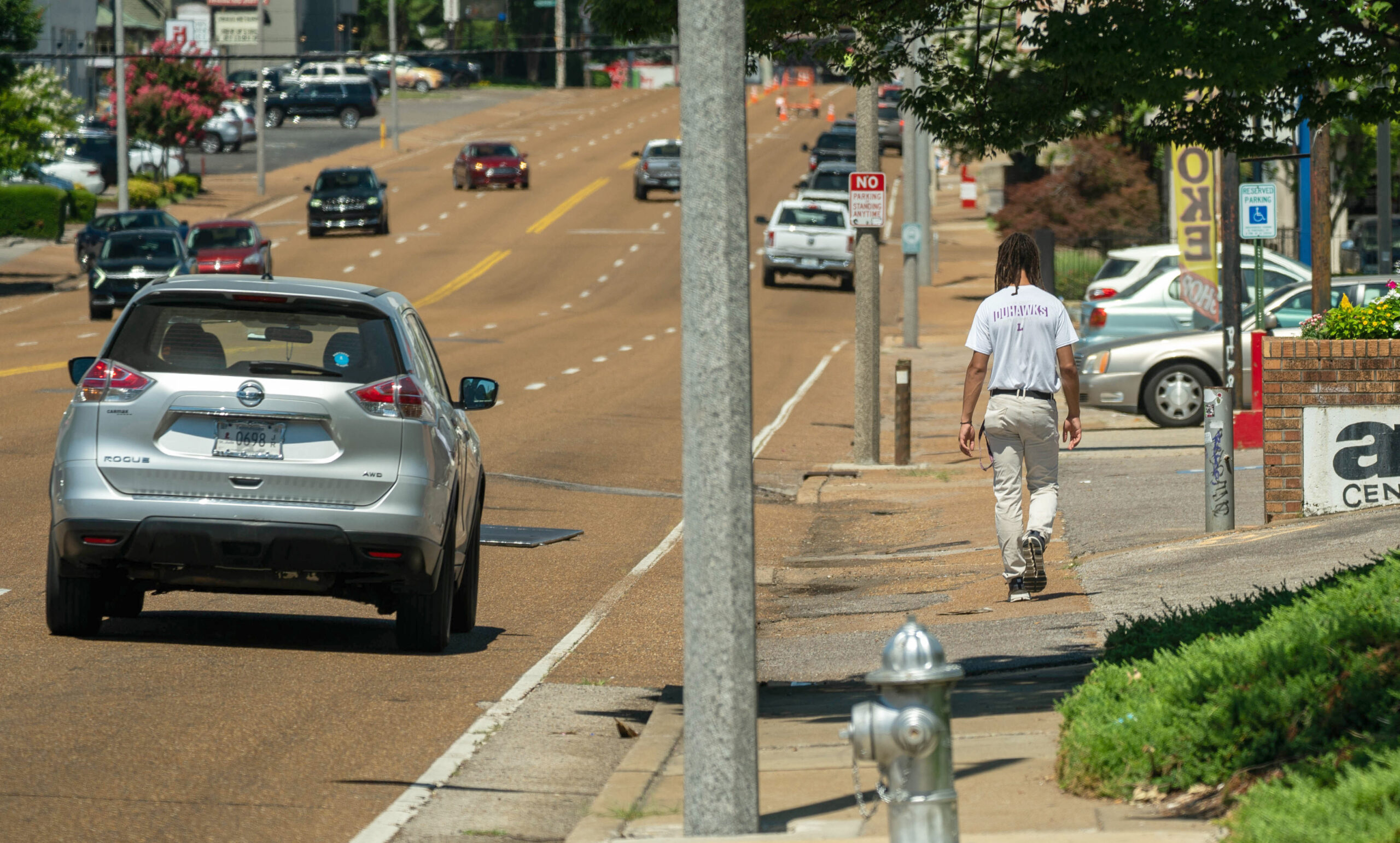 a man walks along a sidewalk with many curb cuts next to six lanes of arterial road traffic