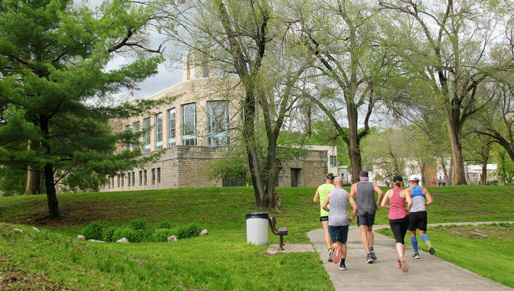 Joggers running with their backs facing on a train through green grass and spring trees with a building in the background