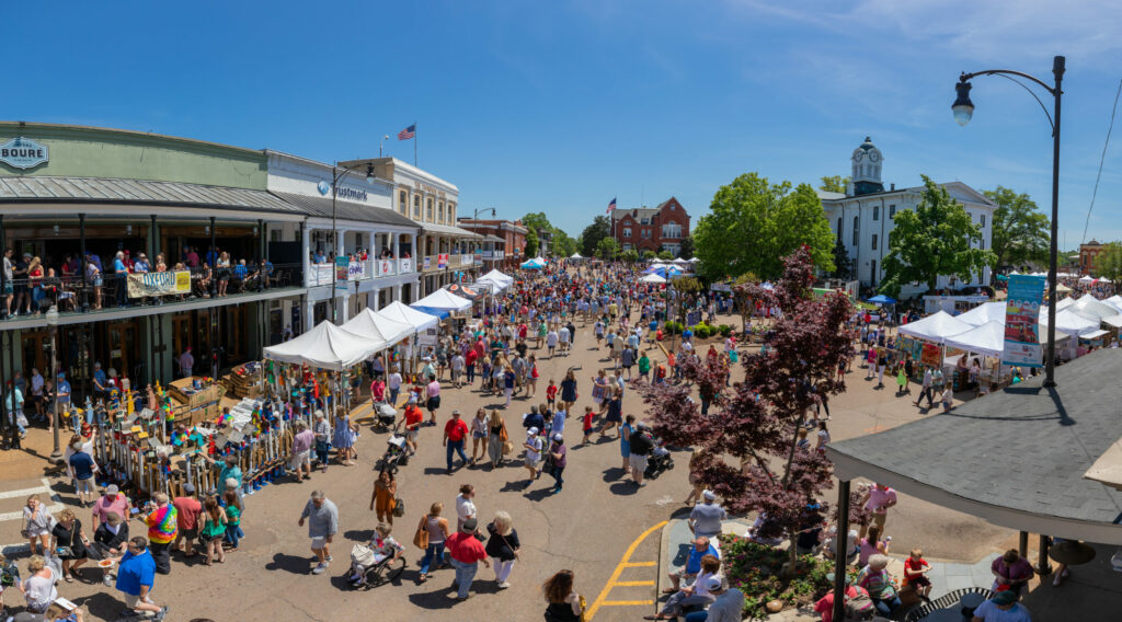 A wide shot of people using the street, with a street fair happening and buildings with second floor patios filled with people.