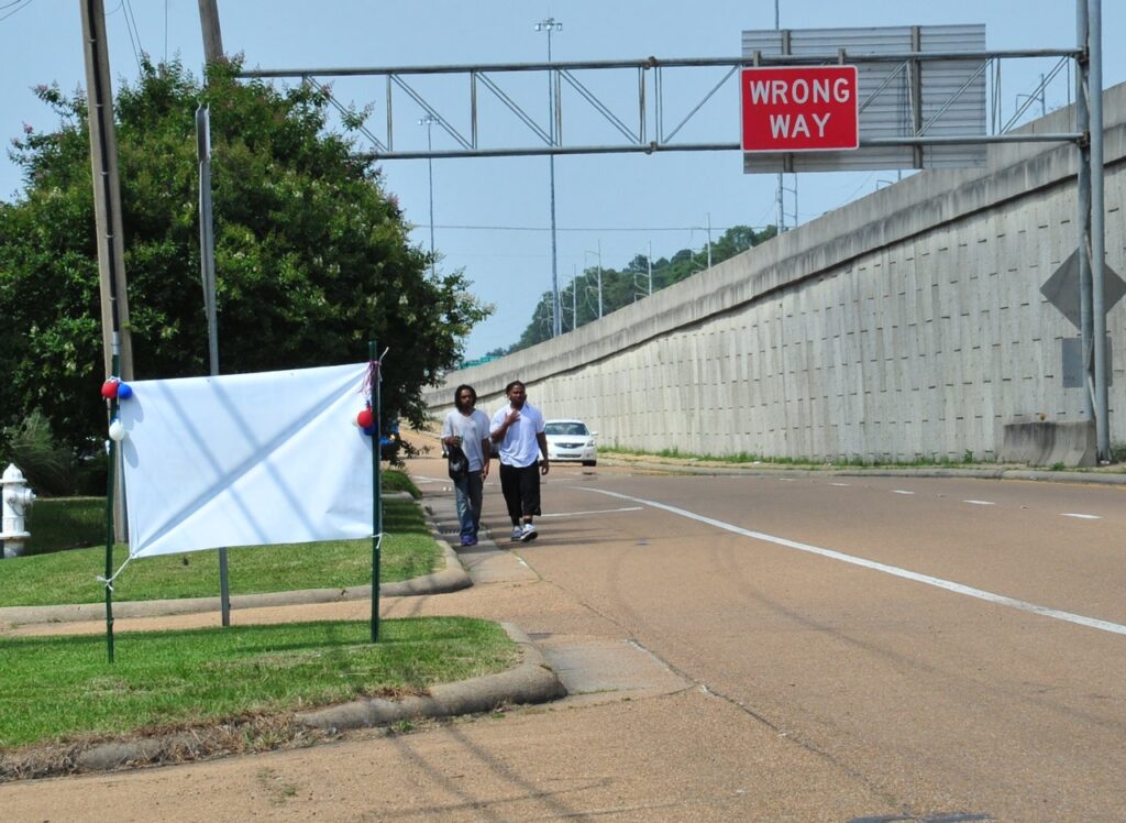 Two people walk in the street lacking sidewalks next to the bare wall of an elevated interstate