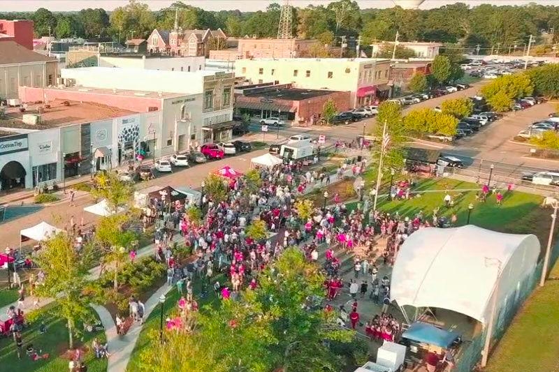A small park with a tent that appears to be covering a stage. There are many people in front of it, many of who are wearing bright pink shirts. A small downtown is in the background