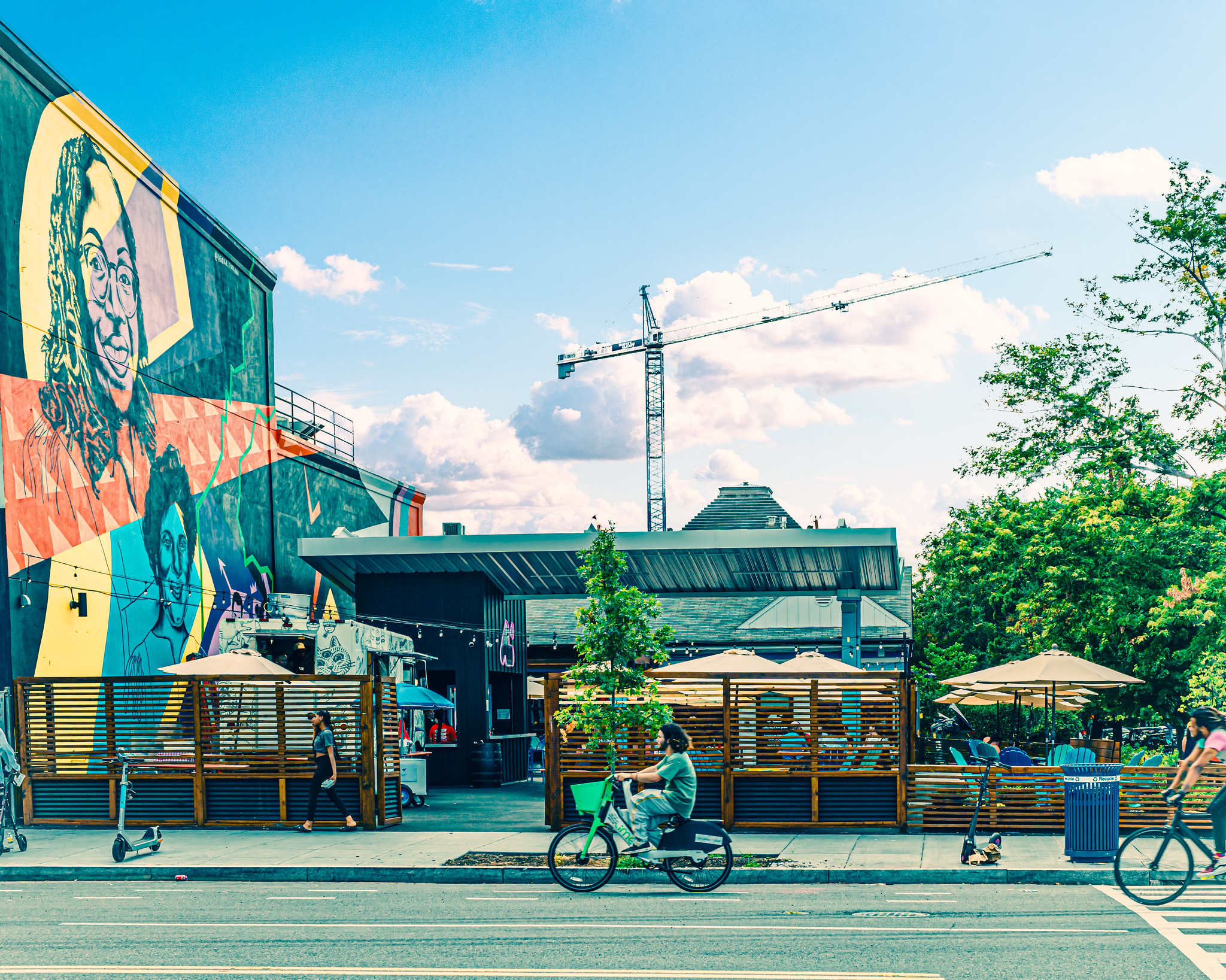 A cyclist travels down a bike lane on DC's 14th street. In the background is a sidewalk with parked scooters, patio seating for a local business, and a colorful mural.