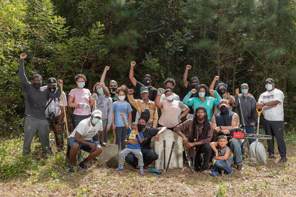 A group of people in the a forested area with shovels, saws, and other working tools with their fists raised. 