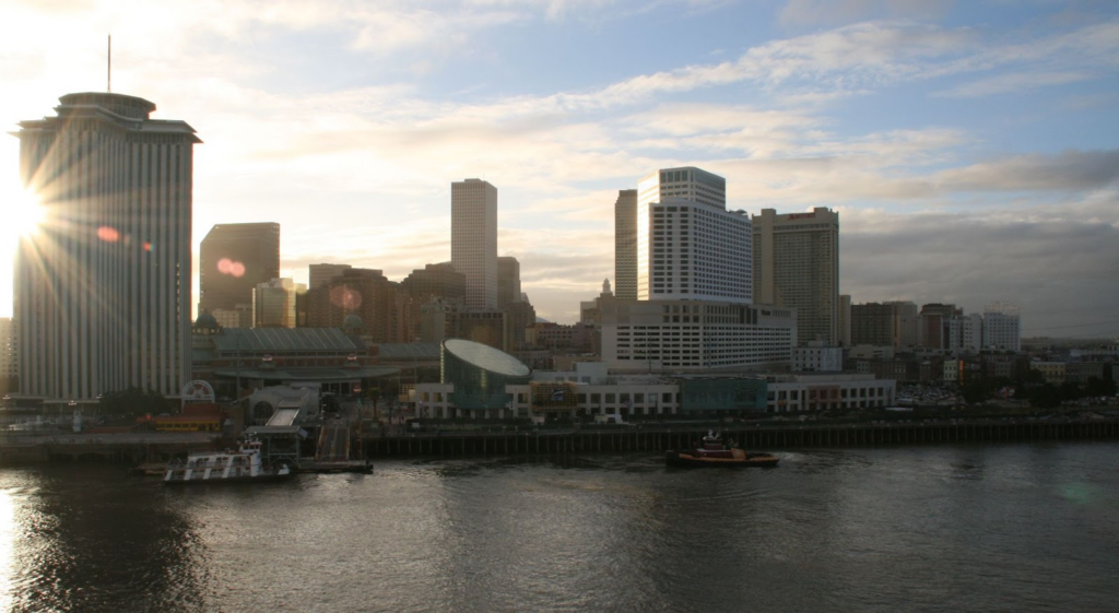 An image of the New Orleans skyline, taken from the water, with the sun shining from behind the buildings