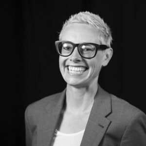 Black and white image of a woman with short, light-colored hair with thick, black-rimmed glasses and a suit jacket, smiling broadly