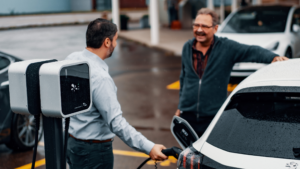 Two men stand, chatting, beside a car while it's getting plugged in to charge.