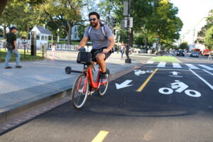 A man with dark hair and sunglasses rides a Capital Bikeshare bicycle down a painted bike lane near a treelined sidewalk.