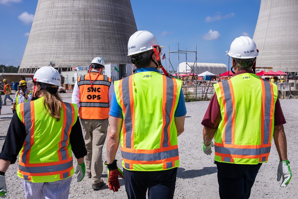 A group of people in yellow and orange safety gear and hard hats walking toward a Nuclear cooling tower
