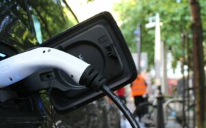 A black EV charges on the side of a tree-lined street. In the background, a construction crew in orange vests mingles on a wide sidewalk with bike parking.