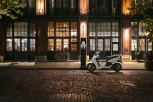 An electric scooter charges at the curb in front of a warmly lit storefront at night