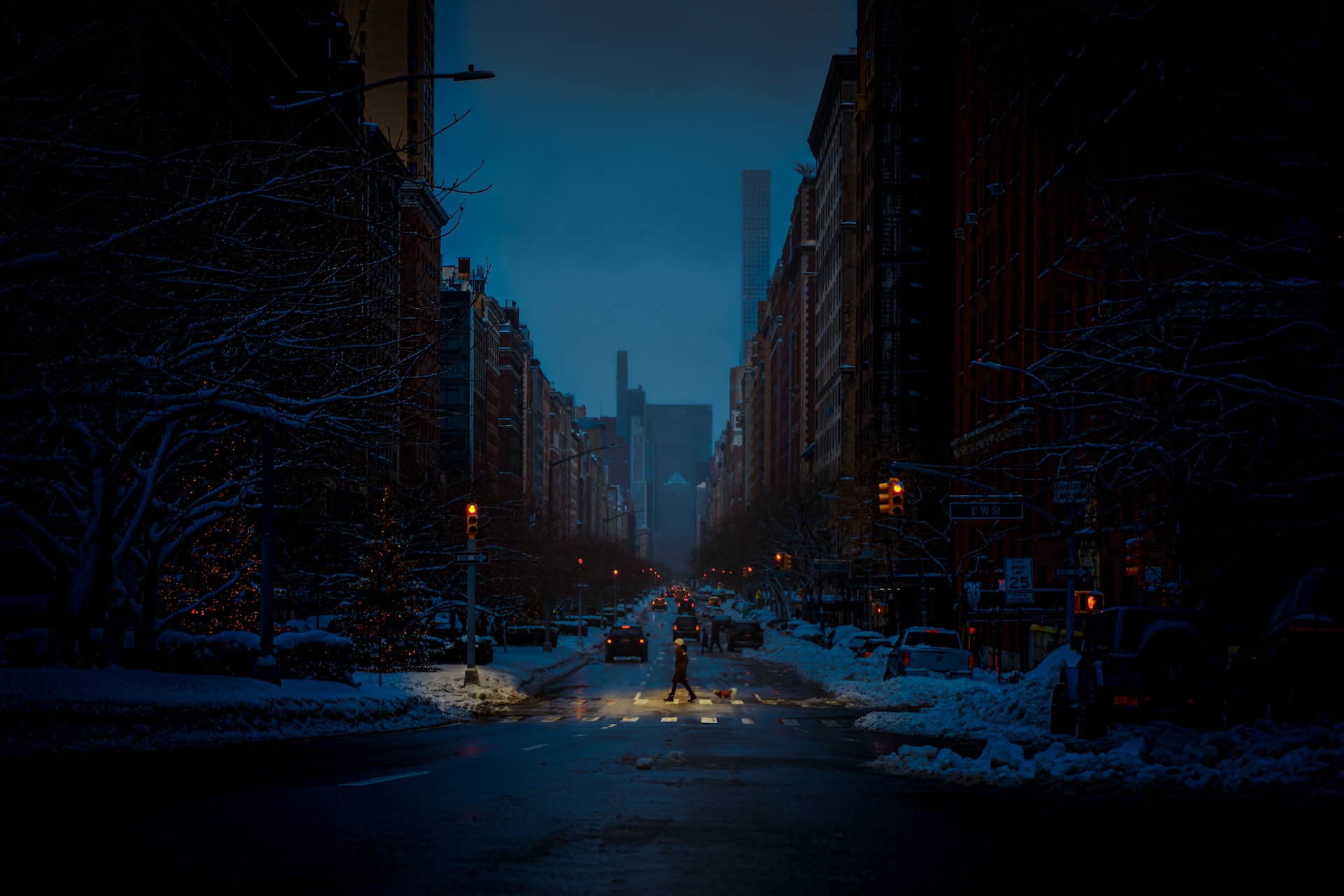 Photograph of a dark street with snow built up on the sides of the road and cars approaching in the background. A single pedestrian is crossing the street, dimly illuminated by a lone streetlight.