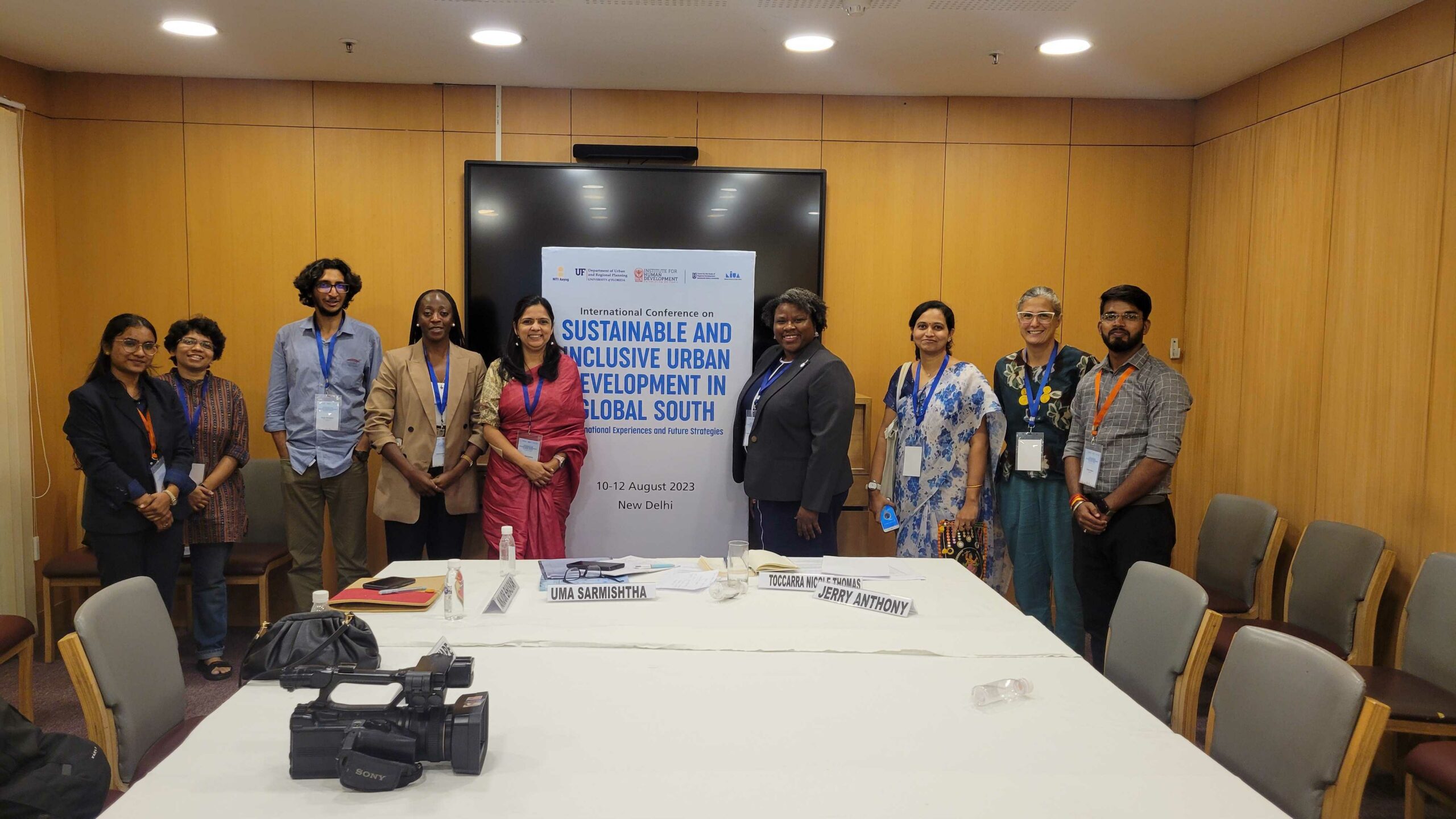 A group of people stand around a poster that says sustainable and inclusive urban development in the global south august 10th through 12th 2023 New Delhi