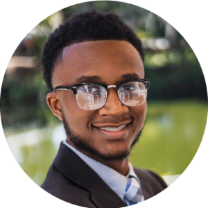 A portrait of Isaiah wearing glasses, a blue buttoned up shirt and suit coat and tie.