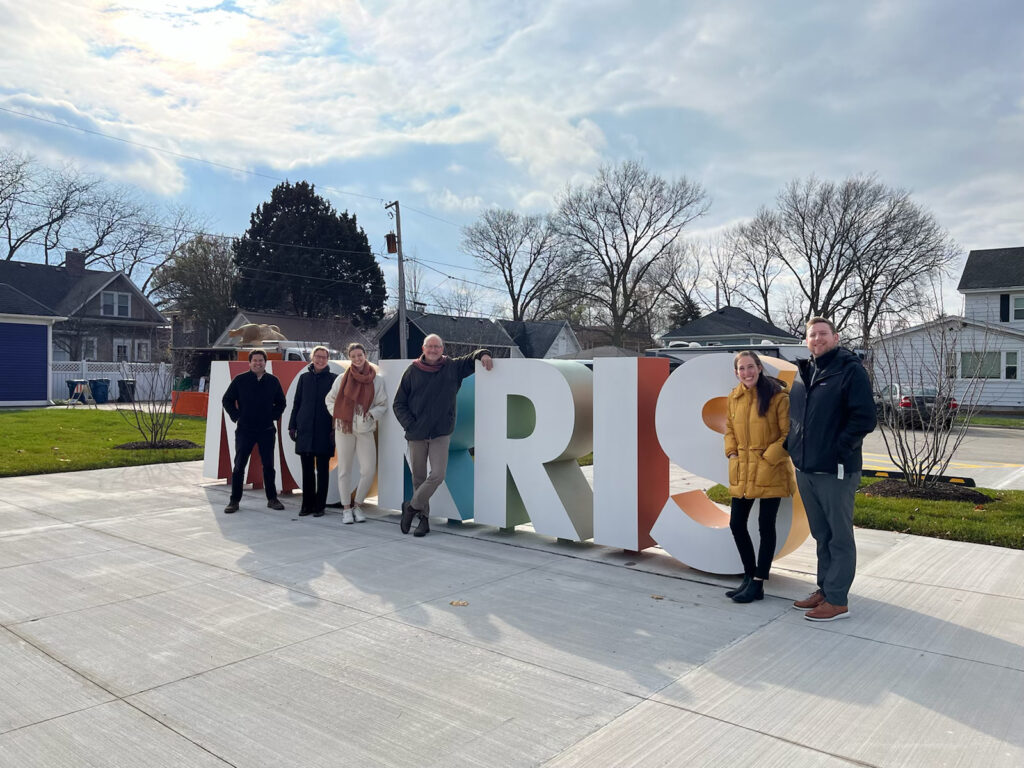 Six employees in front of colorful text sculpture that reads "Morris" during what looks to be a chilly day. Trees in the background have no leaves 
