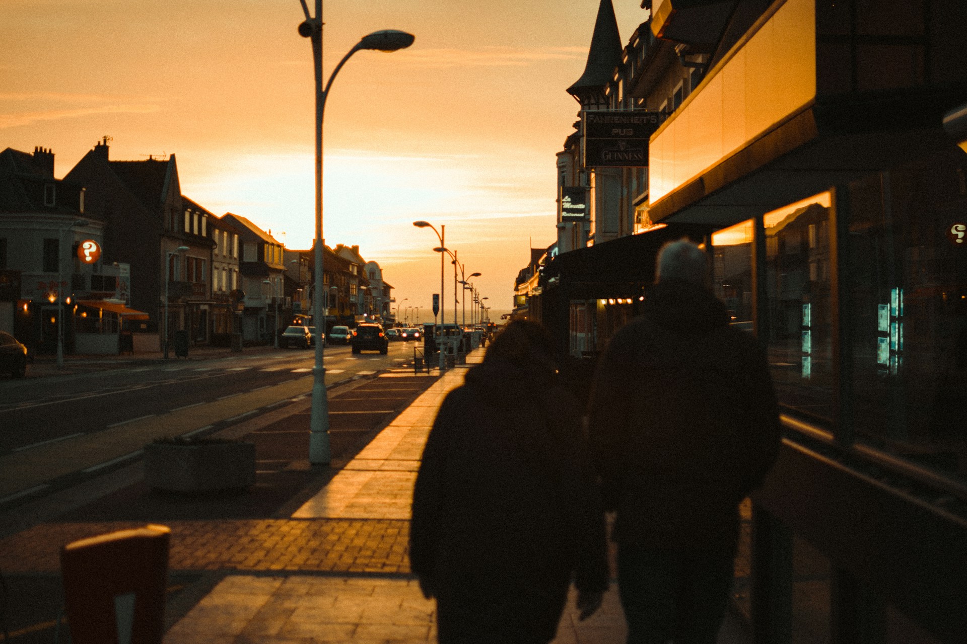 An elderly couple in winter coats walks down a wide sidewalk next to a calm roadway beside shops and a church
