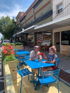 A couple clinks glasses in the new parklet dining space, surrounded by flowers and streetside shops