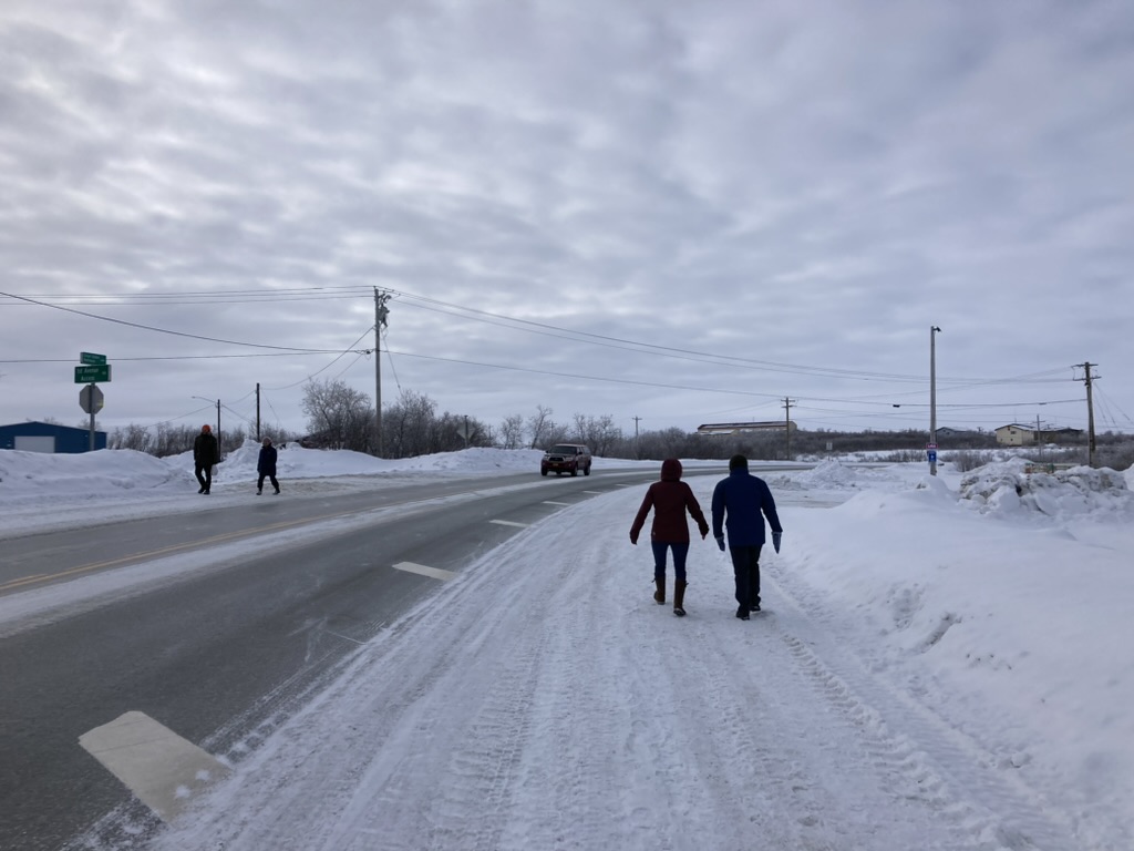 Two people walk on the snow off the side of the road. 