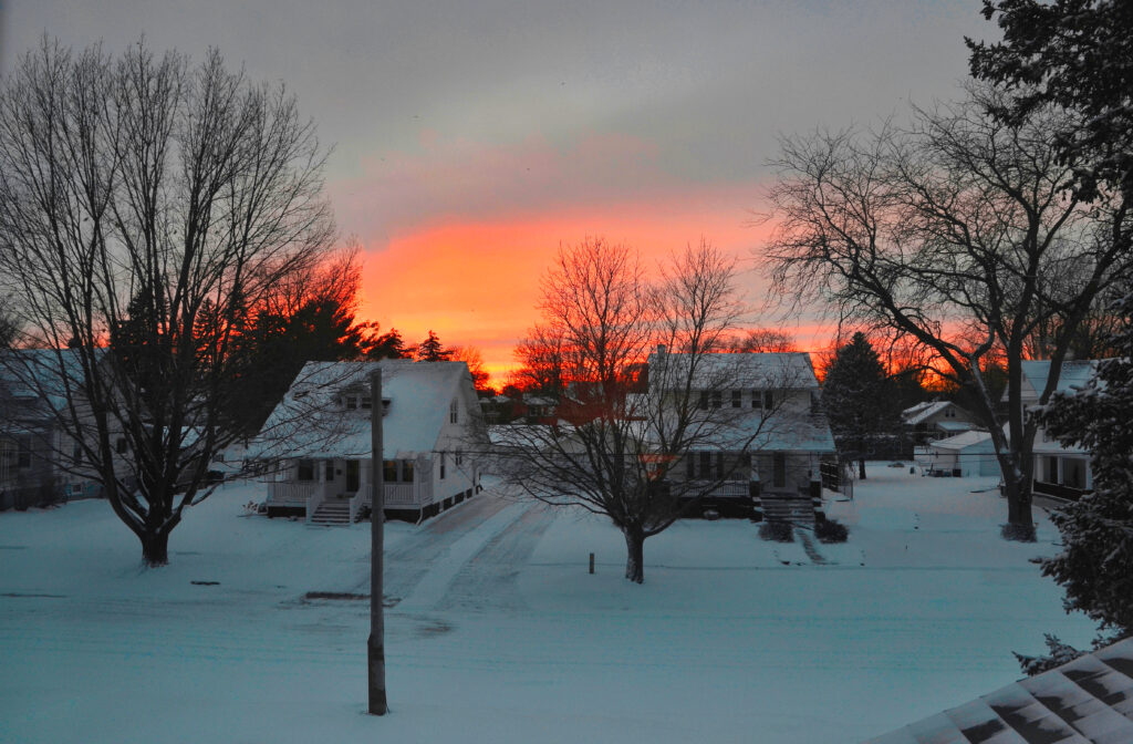The sunsets behind two houses in a neighborhood covered in snow. 