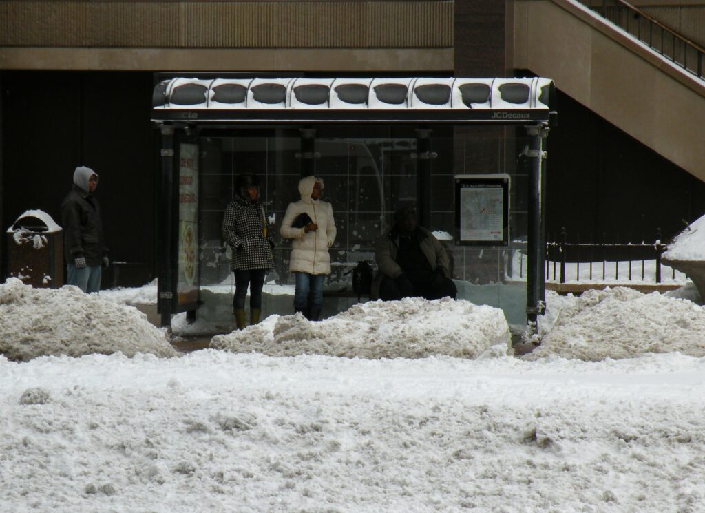 People stand under a covered bus stop where snow is piled up from being plowed.